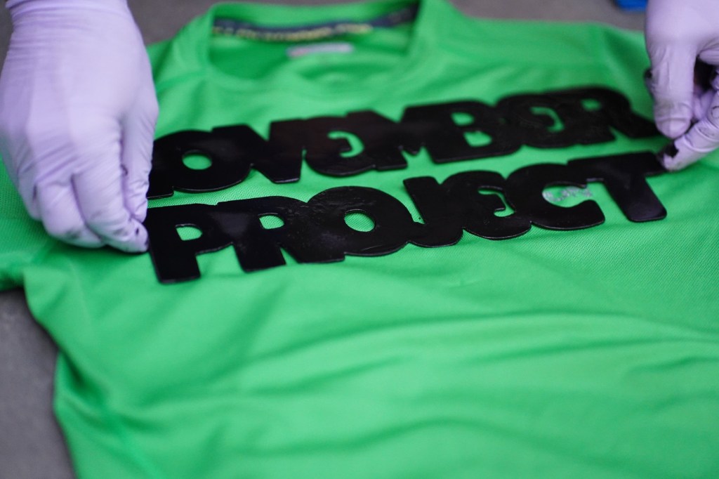Image of a green t-shirt in the process of being tagged with the November Project logo